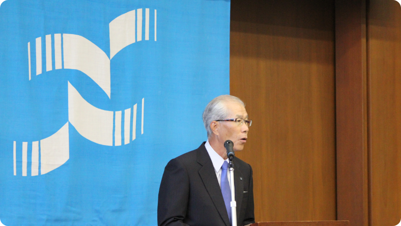 合併式　新しい社旗の前であいさつする芳賀社長